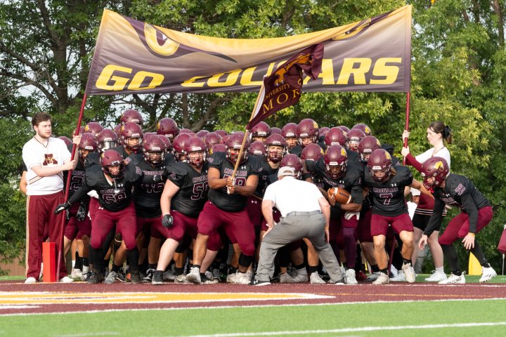 A football team, dressed in maroon and black uniforms, prepares to charge onto the field. They stand closely together, led by a coach and a player holding a maroon flag with the team's logo. Above them, a large banner reads 'GO COUGARS,' as a cheerleader holds one side of it. 